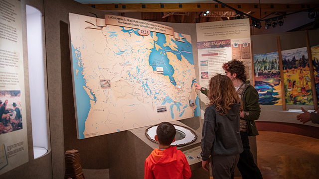 Deux adolescents et une jeune garçon sont de dos; ils regardent un panneau d’interprétation de l’exposition Obadjiwan présentée au lieu historique national d’Obadjiwan–Fort-Témiscamingue.