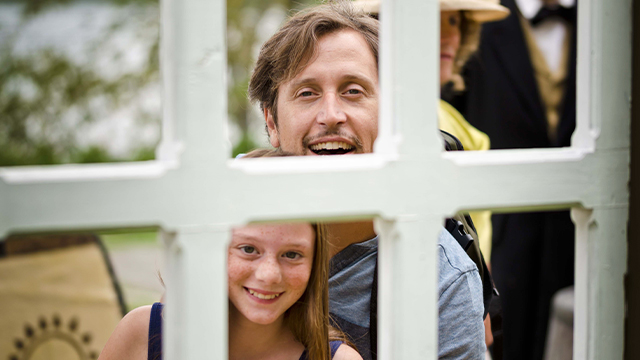 Un papa et sa fille sourient et regardent à travers les carreaux d’une fenêtre d’une des scénographies extérieures au lieu historique national d’Obadjiwan–Fort-Témiscamingue.