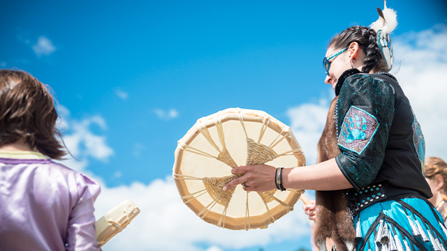 Une femme joue du tambour autochtone traditionnel aux côtés de fillette au lieu historique national d’Obadjiwan–Fort-Témiscamingue lors d’une superbe journée d’été où le ciel est très bleu.
