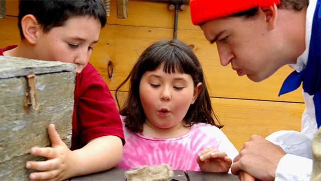 Deux enfants, un garçon et une fille, en compagnie d’un guide costumé soufflent sur de la fourrure lors d’une visite scolaire au Lieu historique national d’Obadjiwan–Fort-Témiscamingue.