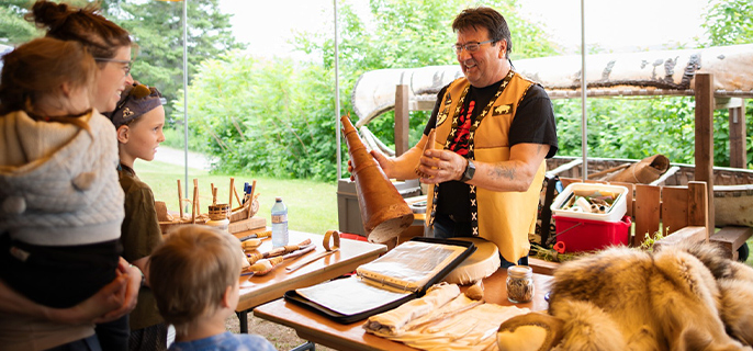 L’artiste contemporain et artisan Anichinabé, Karl Chevrier, est sous un chapiteau au lieu historique national d’Obadjiwan–Fort-Témiscamingue; il montre à une femme et ses trois jeunes enfants le cornet d’appel à l’orignal qu’il a fabriqué.