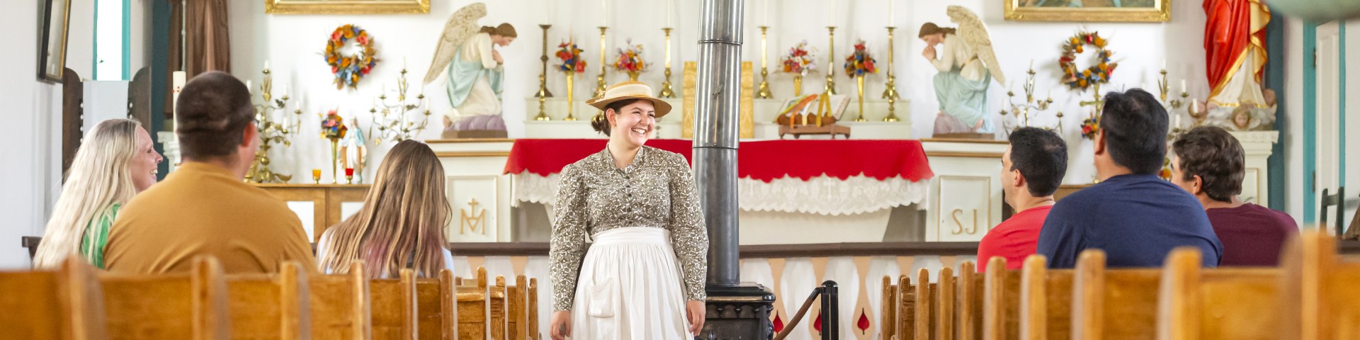 A Parks Canada interpreters stands in front of visitors seated in the St. Antoine de Padoue church.