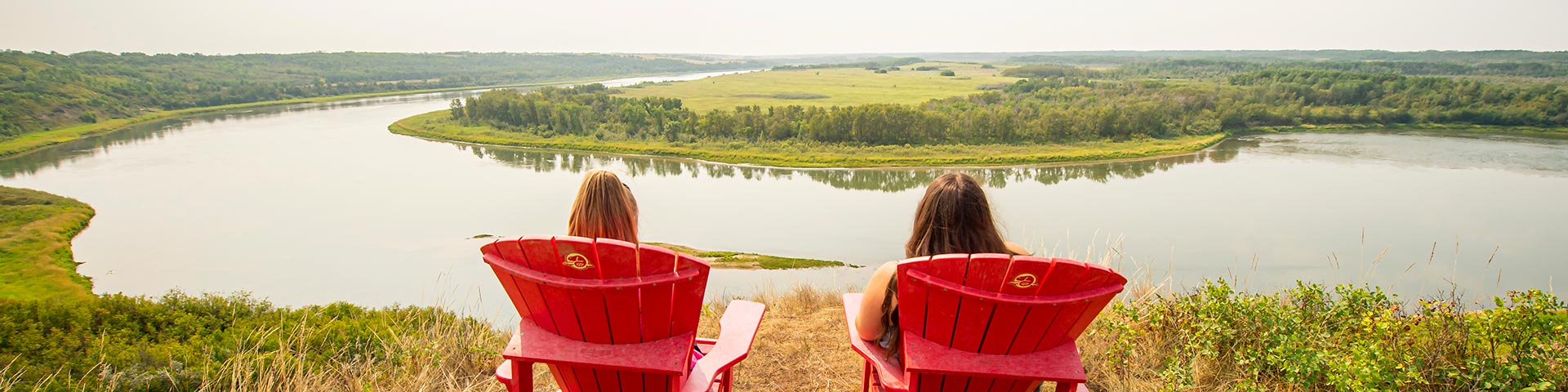 Two visitors sit Parks Canada red chairs overlooking the river. 