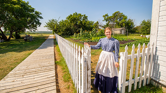 A woman in historic costume standing outside a white building near a boardwalk, with a garden in the background