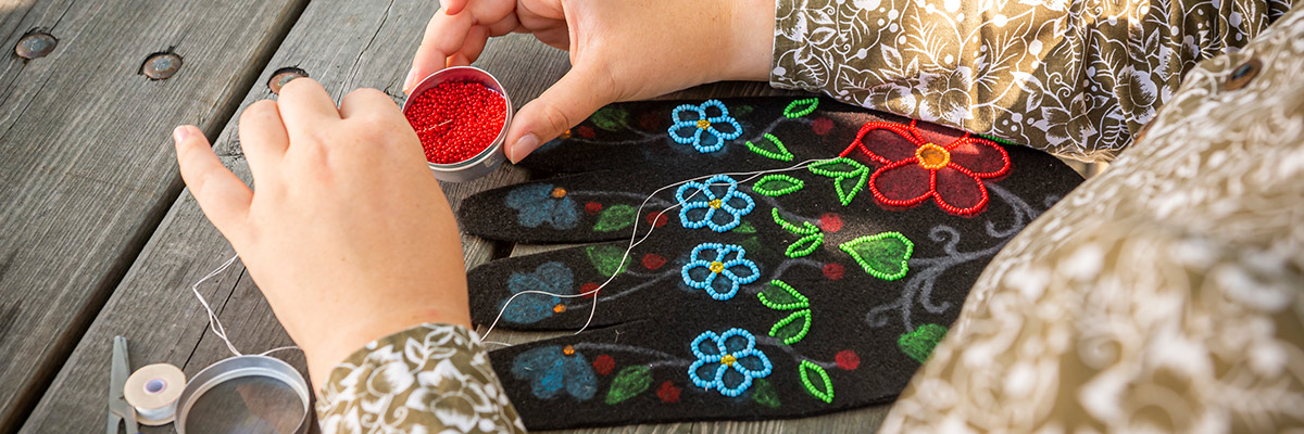 A person sitting at a picnic table stringing red beads overtop of a piece of material with beadwork on it.