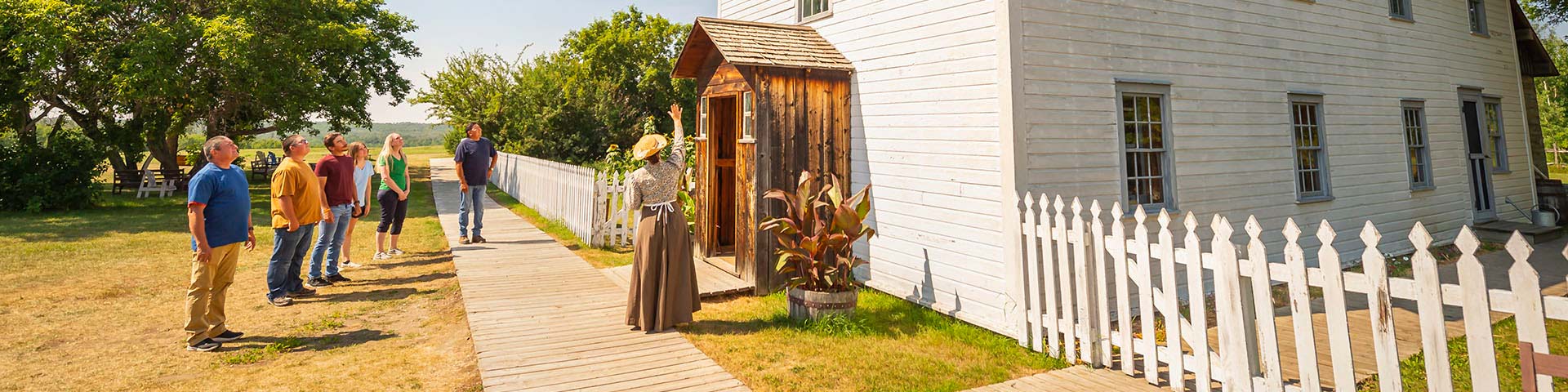 A Parks Canada interpreter points up to the bullet holes in the Rectory while a group of visitors stand in front.