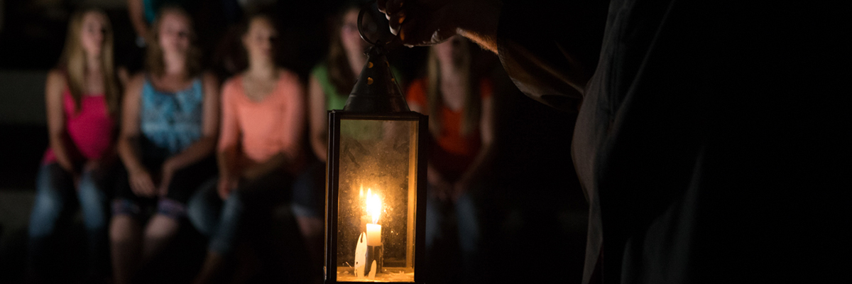 A lit lantern held by the hand of a cloaked figure in front of a group of visitors. 