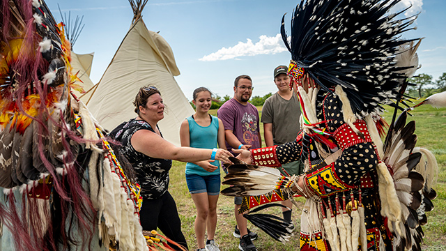 Two Indigenous presenters in traditional regalia speak to a group of visitors. 