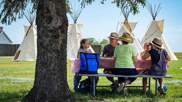 Two adults and three children enjoy a picnic at a shaded picnic table with tipis in the background. 