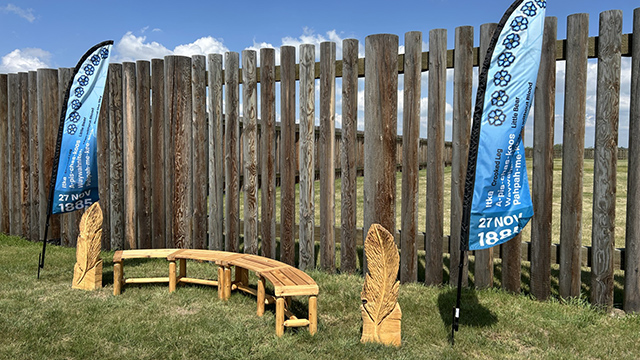 A wooden bench with two wood feather carvings and blue flags with the names of the eight men executed on November 27, 1885.