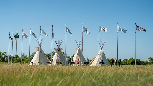 Twelve flags ripple in the breeze in front of four tipis as visitors view interpretive panels. 