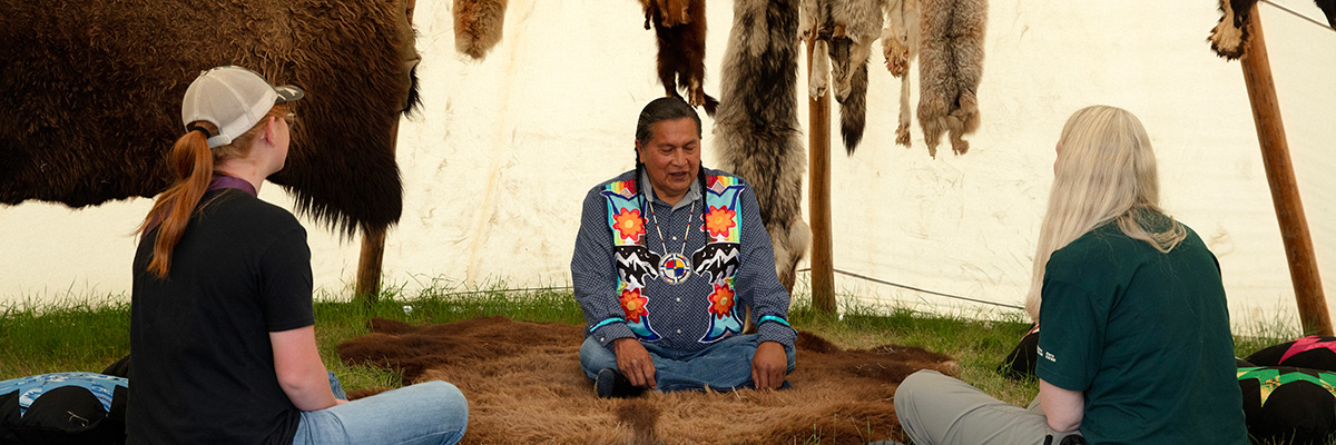 Indigenous presenter wearing a ribbon shirt inside a tipi speaking to visitors. 