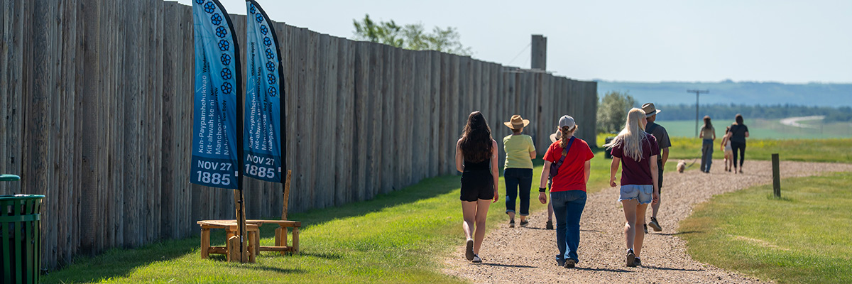 Visitors walk along the pathway outside the palisade past a wooden bench with two blue flags.