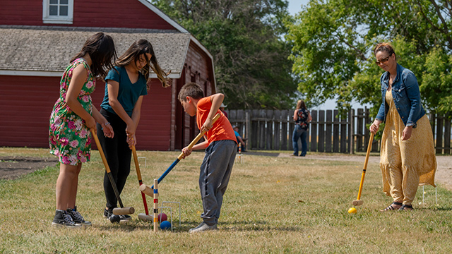 A family playing croquet with a red building in the background.