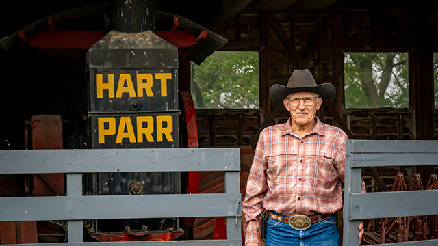 A man in a cowboy hat stands by a gate in front of a Hart Parr tractor in a shed.