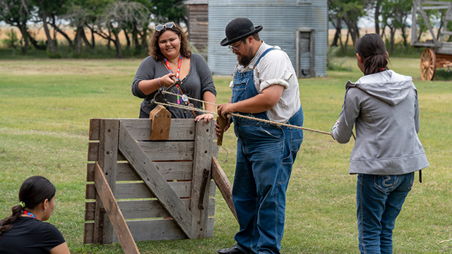 Two women hold the ends of a rope while a man in period costume twists it together in the middle.