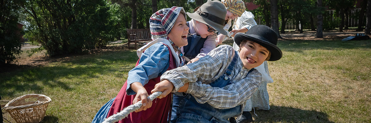 children in historic clothing pull on a rope for a game of tug-of-war.