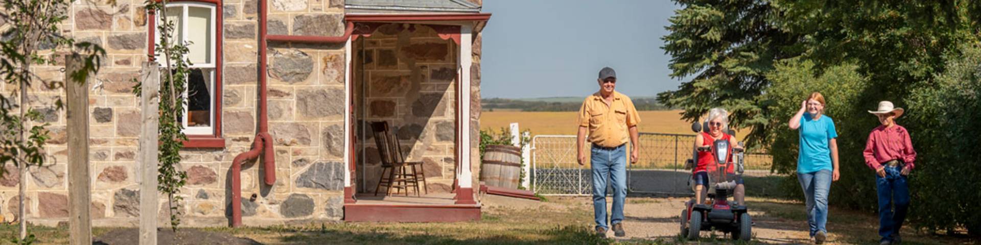 An adult visitor on an electric scooter with an adult and two children walking on a pathway near the stone house in summer at Motherwell Homestead National Historic Site. 