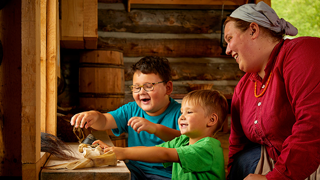 Deux jeunes garçons essaient des outils fabriqués à partir de bison avec une femme en costume d’époque.