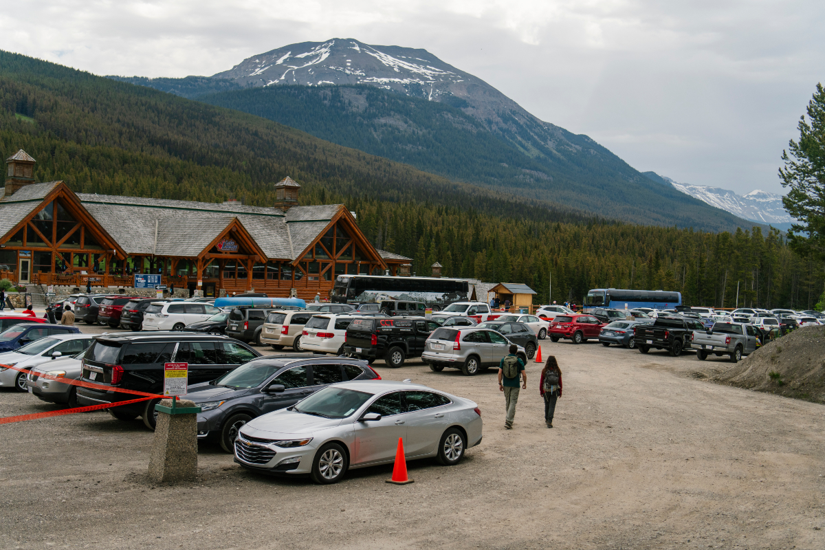 A parking lot in the foreground with a wooden building, treed slopes, and mountain in the background. 