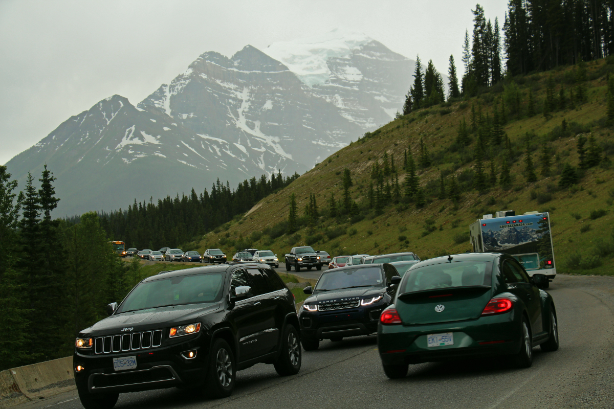 Vehicles travel bumper to bumper along a two-lane roadway with a grassy hill on one side and a mountain in the distance.    