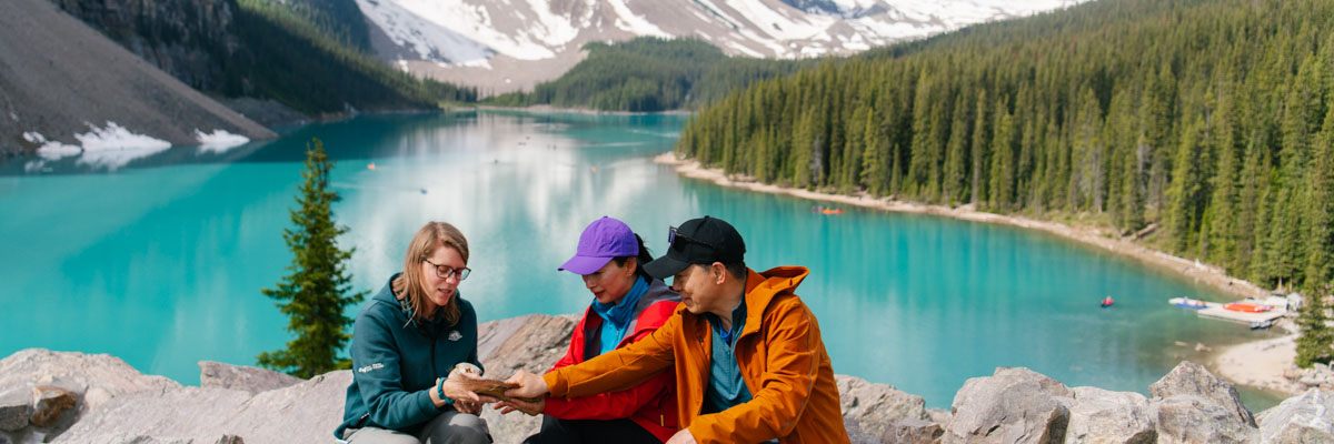 A Parks Canada interpreter shows details of a rock to two visitors. They are sitting on rocks facing the camera with Moraine Lake and mountains behind them