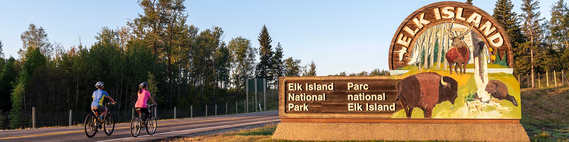 Des cyclistes passent à vélo devant le panneau d’entrée de la porte nord du parc national Elk Island