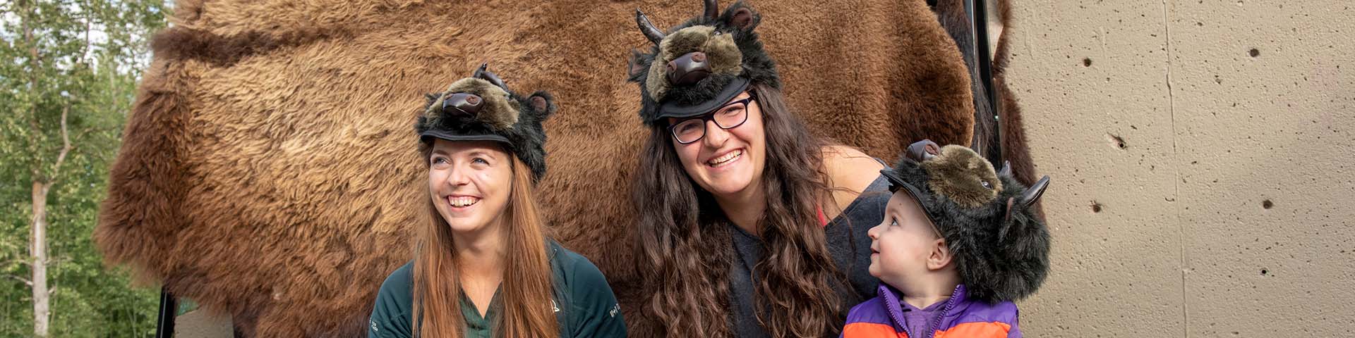 Une mère et une fille essaient des chapeaux en fourrure de bison avec un interprète de Parcs Canada au théâtre Astotin dans le parc national Elk Island. Parc national Elk Island