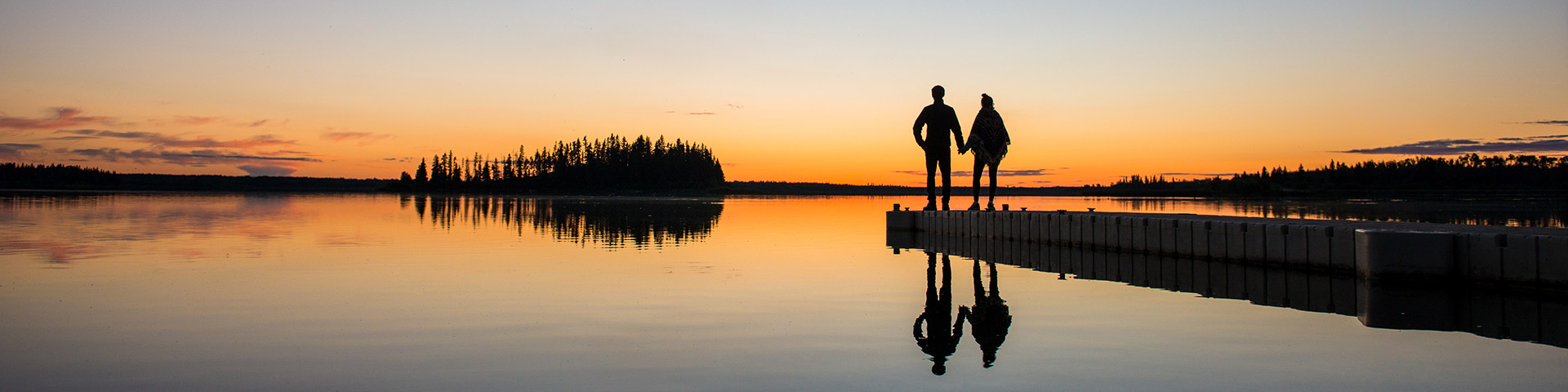 A couple a silhouetted while standing on a dock watching a sunset at Astotin Lake in Elk Island National Park.