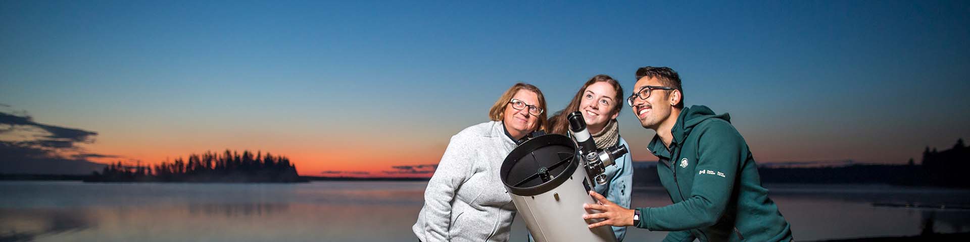 Un interprète de Parcs Canada à côté d’un télescope discute avec des visiteurs pendant un coucher de soleil au lac Astotin, dans le parc national Elk Island