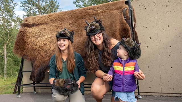 Une mère et une fille essaient des chapeaux en fourrure de bison avec un interprète de Parcs Canada au théâtre Astotin dans le parc national Elk Island. Parc national Elk Island