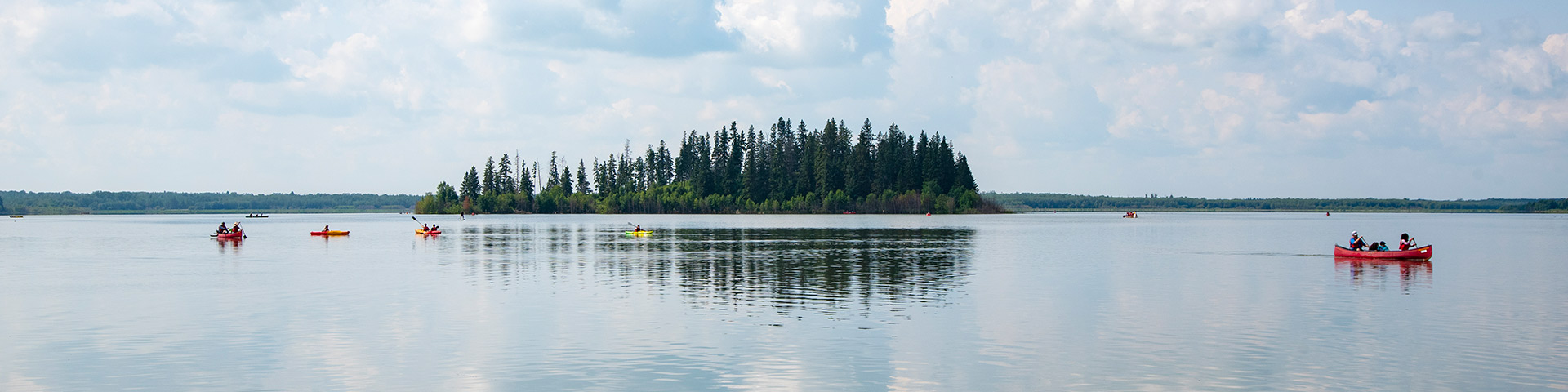 A photo taken from a distance of paddlers enjoying a calm summer day on Astotin Lake. A skyline of trees is visible in the background. 