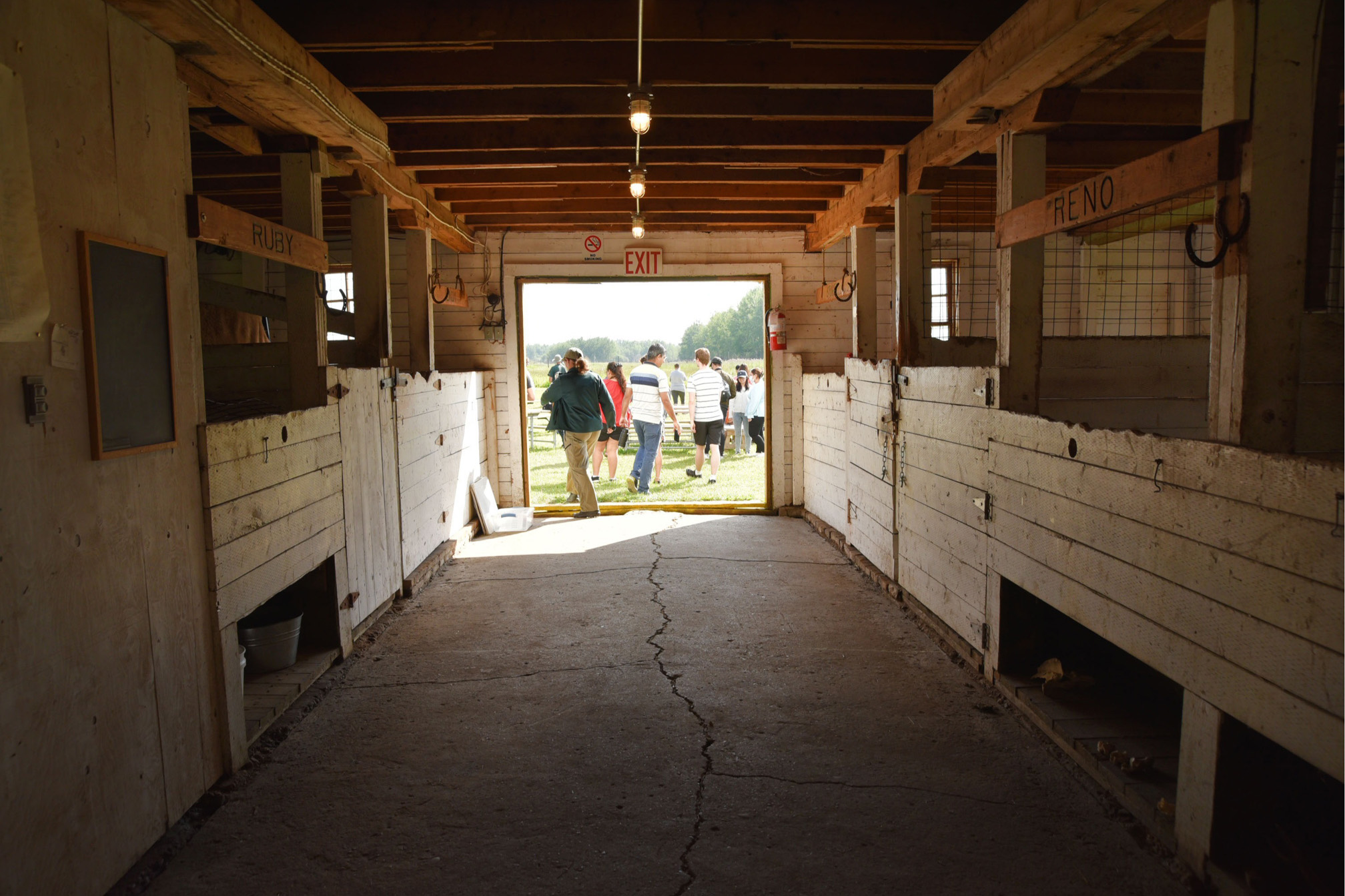 Les stalles de chevaux sont visibles à l’intérieur de l’écurie alors que des visiteurs et un interprète sortent par les portes de l’écurie.