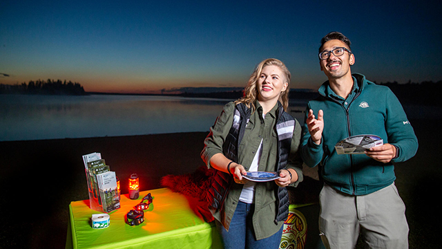 Une interprète de Parcs Canada et une visiteuse regardent le ciel nocturne en tenant des cartes du ciel avec le lac Astotin et une table avec des lampes frontales rouges et des dépliants en arrière-plan.