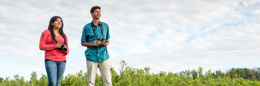 A young couple holding binoculars stand in a meadow. 