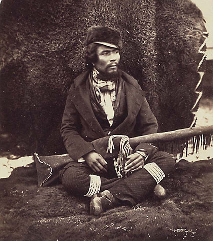 Historical photograph of a man sitting cross-legged on bison hides, holding a gun in a fringed leather bag. 