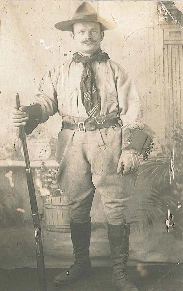 Un homme pose debout dans un studio de portrait, portant un Stetson et un mouchoir de cou. Ses gants, ses bottes et sa culotte servent à monter à cheval.