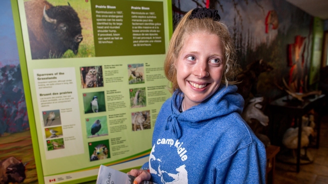 Un visiteur se tient debout et sourit dans l’atrium du théâtre Astotin tout en tenant un Guide du visiteur.
