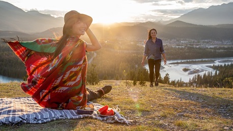 Woman sits on a picnic blanket looking back, another walks toward her; river and mountains behind.