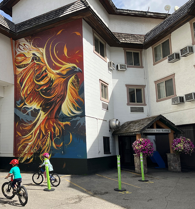 Three-story building with a colorful phoenix rising mural, and two kids riding bikes in front of it.