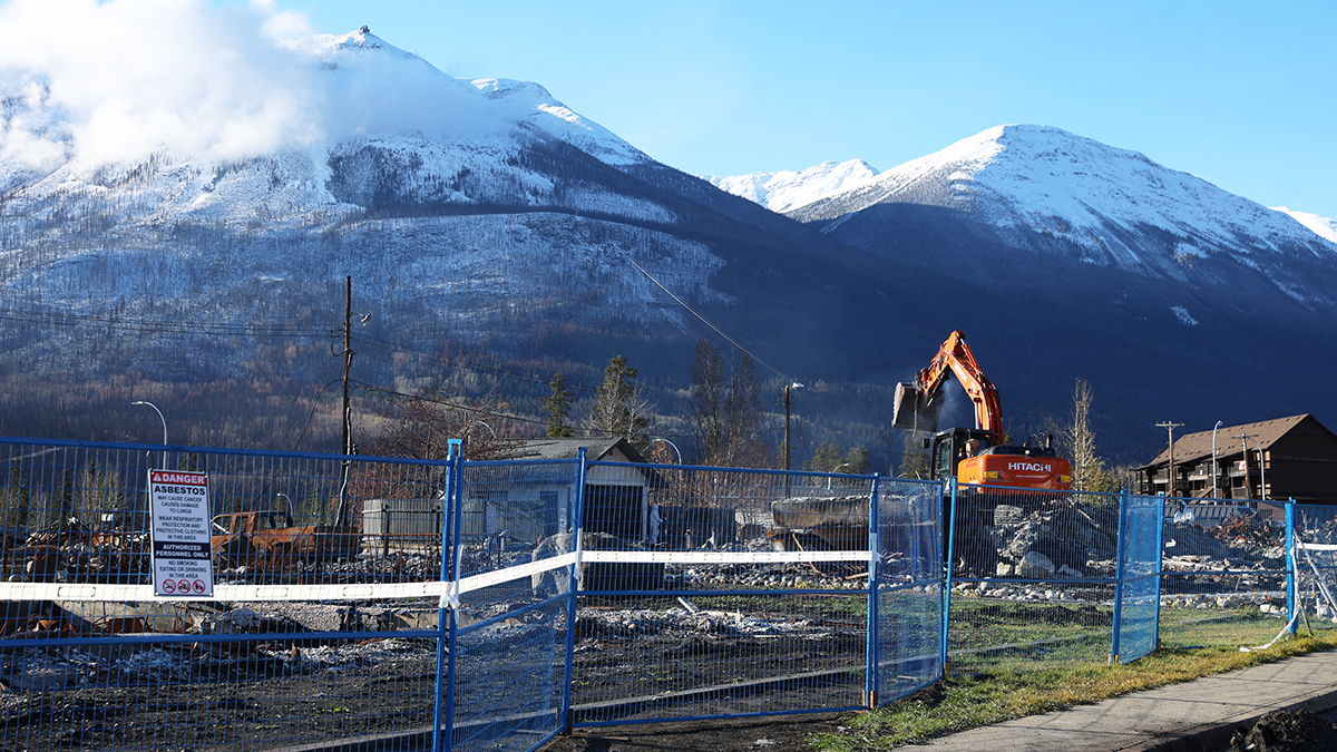View of a debris clearing site with a yellow excavator on the right, blue safety fencing in the foreground, and snow-capped mountains in the background.