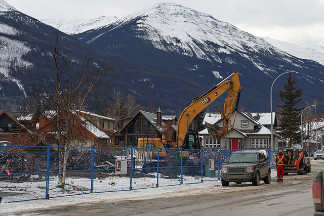 View of a debris removal site with a yellow excavator on the right, blue safety fencing enclosing the area, a black car on the road, and snow-capped mountains in the background.