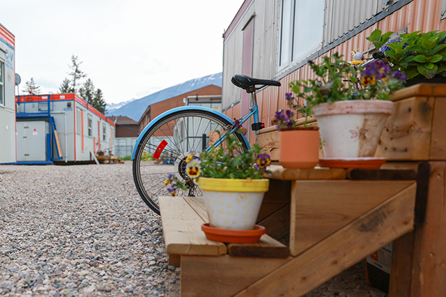 Close-up of an outdoor wooden staircase with plants, a blue bike behind it, and other interim housing units in the background.