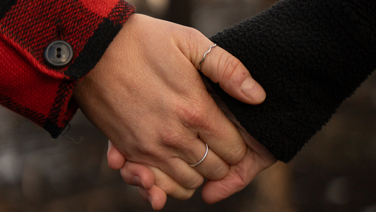 Close-up of two hands holding each other. One hand is wearing a red and black plaid coat and two rings, and the other is wearing a black coat.