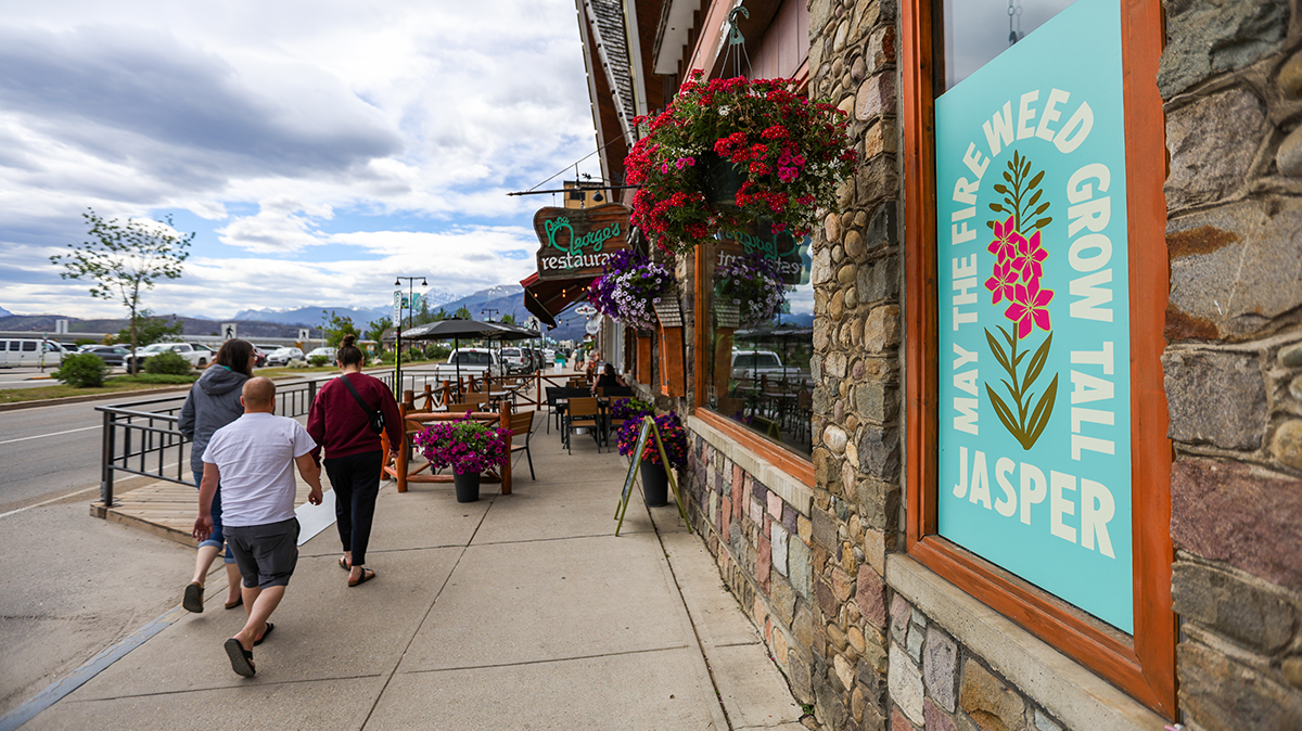 Sidewalk with three people walking away, flowers hanging by a window and a sign in the foreground reading “May the fireweed grow tall Jasper.”