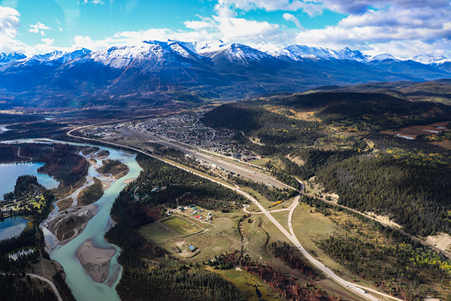 Aerial view of the east side of Jasper townsite after the July 2024 wildfire with snow-capped mountains in the background, a highway, and a river running along the left side.