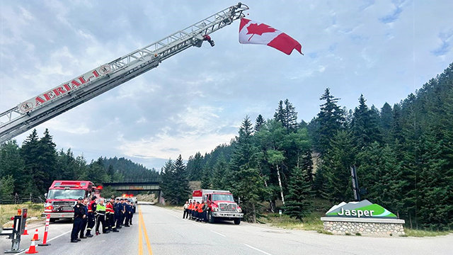 A road with emergency crew members lined up in two rows, with two firefighter trucks behind them. The Jasper town sign is to the right, a large crane is over the group of people with a Canadian flag flying overhead, and some green trees in the background.