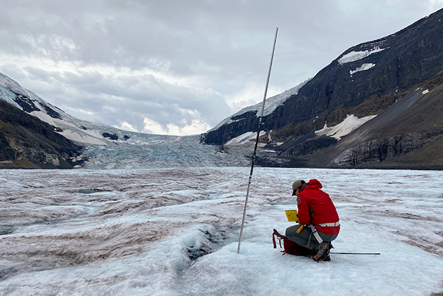 A glacial landscape with a researcher kneeling and examining some equipment, with a long instrument drilled into the ground.