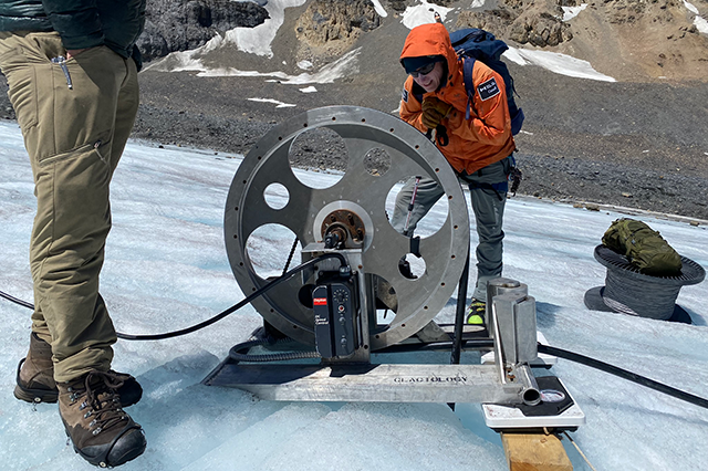 Two researchers, one mostly out of frame, with the other looking directly at a drill on the glacier surface.