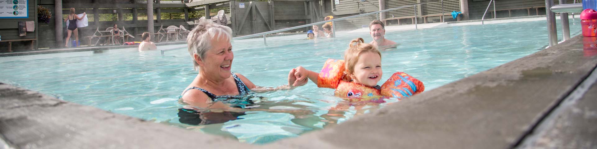 A grandmother and her grandchild playing in Miette Hot Springs on a sunny day, surrounded by large green pine trees in the back drop.